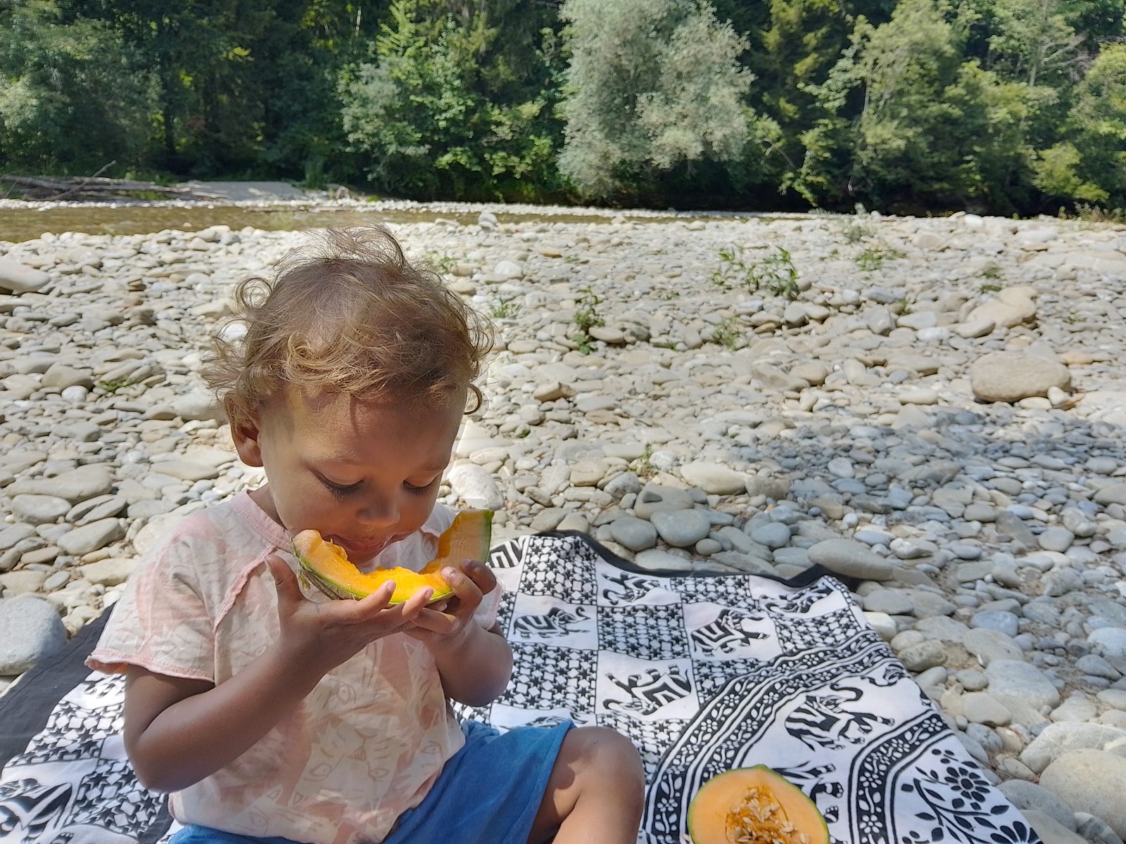Ein Kleinkind isst genüsslich ein Stück Melone am Kiesufer eines Flusses